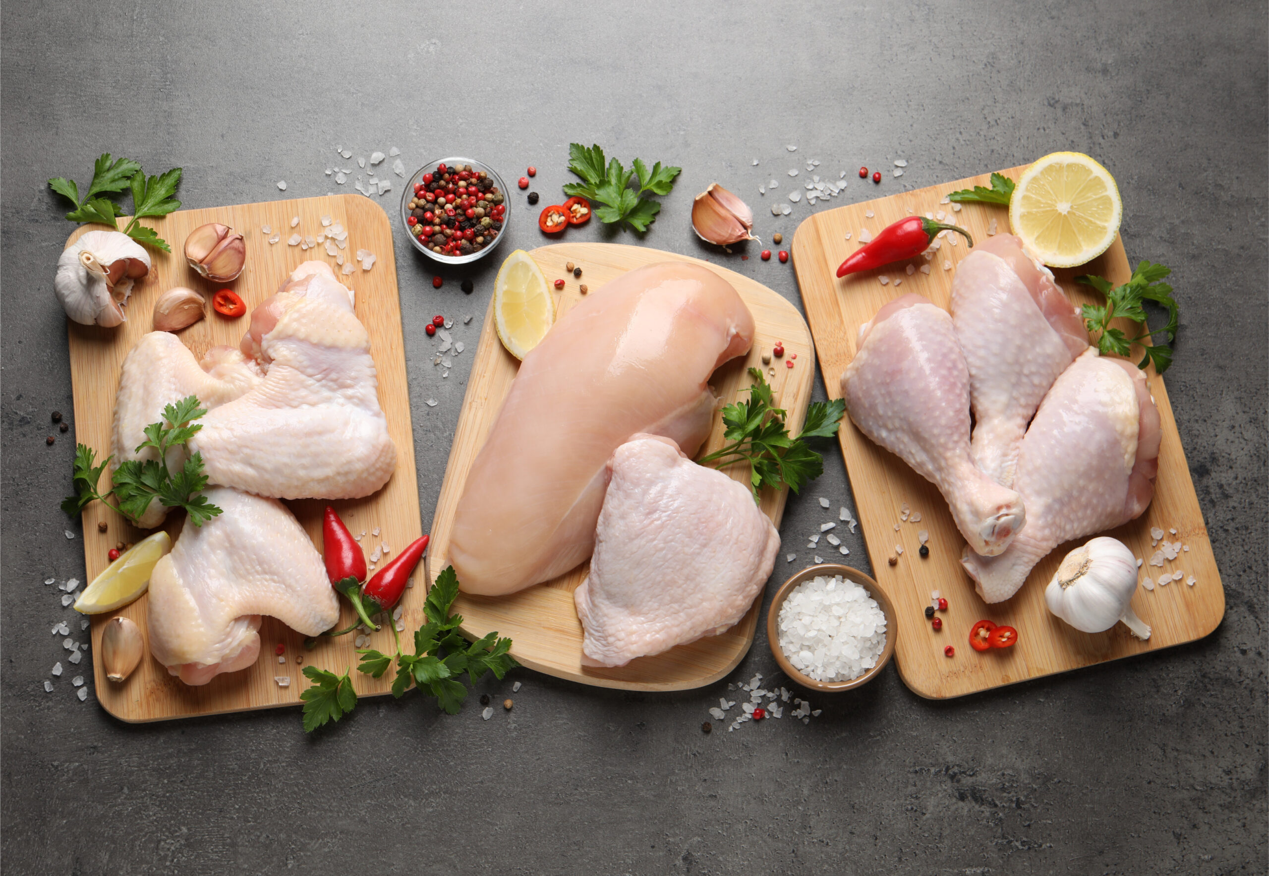 Chicken_Cuts_Assortment_Raw_Breast_Thighs_Drumsticks_Wings_L Fresh chicken cuts displayed on wooden cutting boards, including chicken breast, thighs, and drumsticks, garnished with herbs, garlic, chili peppers, lemon slices, and coarse salt. Ideal for meal preparation and cooking inspiration.